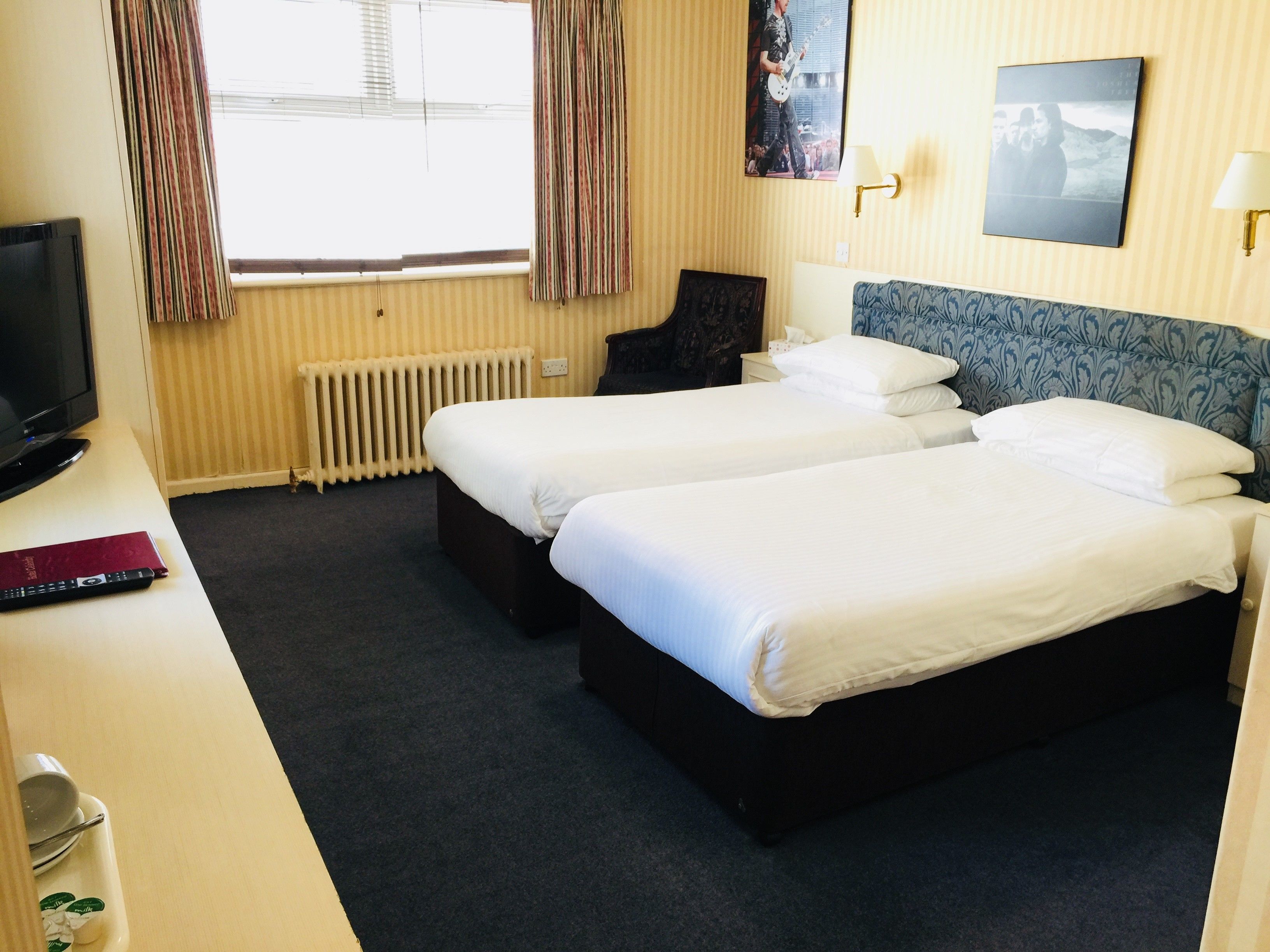 Une chambre d’hôtel avec deux lits, un bureau avec une télévision, un radiateur et une fenêtre avec des rideaux.