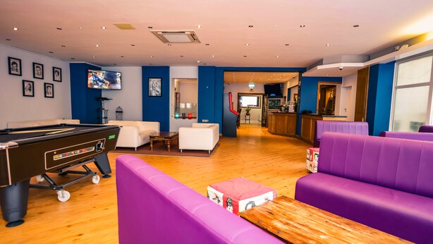 Ein Billardtisch in einem Raum mit violetten Sitzgelegenheiten, einer Bar und einem Fernsehbildschirm.