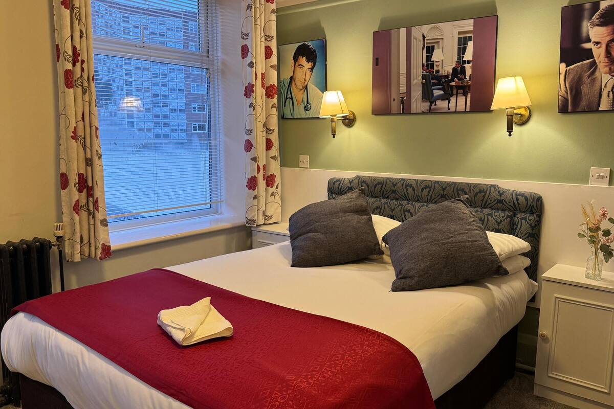 Hotellihuone, jossa on sänky, punainen peitto, kaksi tyynyä, yöpöytä lampun kanssa, maljakko kukkineen ja ikkuna, jossa on verhot.