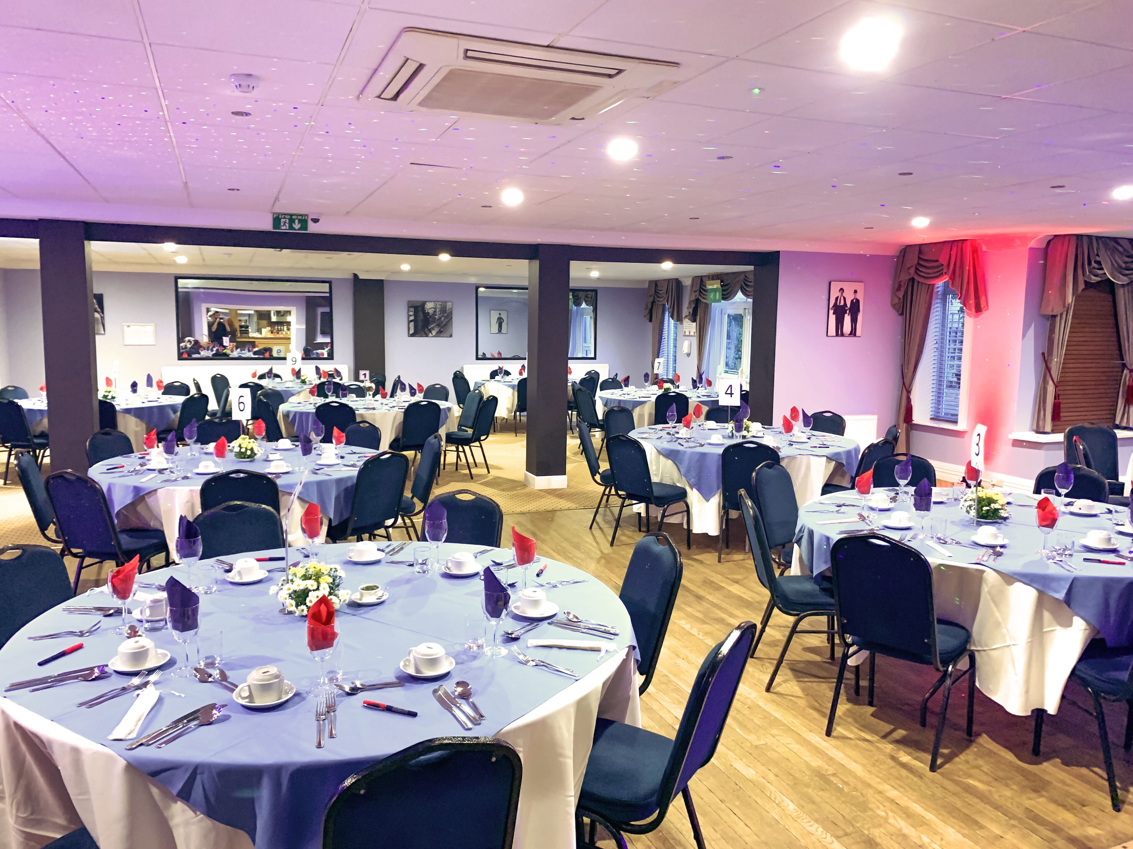 A banquet hall with round tables set for dining, numbered signs, and a view of the kitchen area in the background.