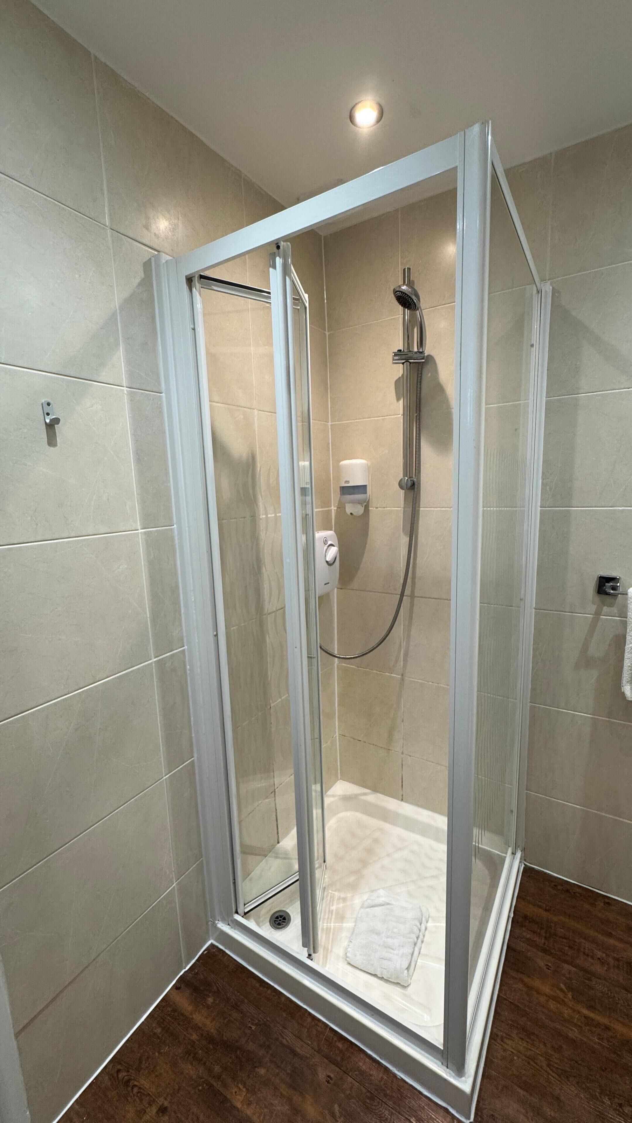 A glass-enclosed shower stall with a handheld showerhead, a soap dispenser, and a towel rack.