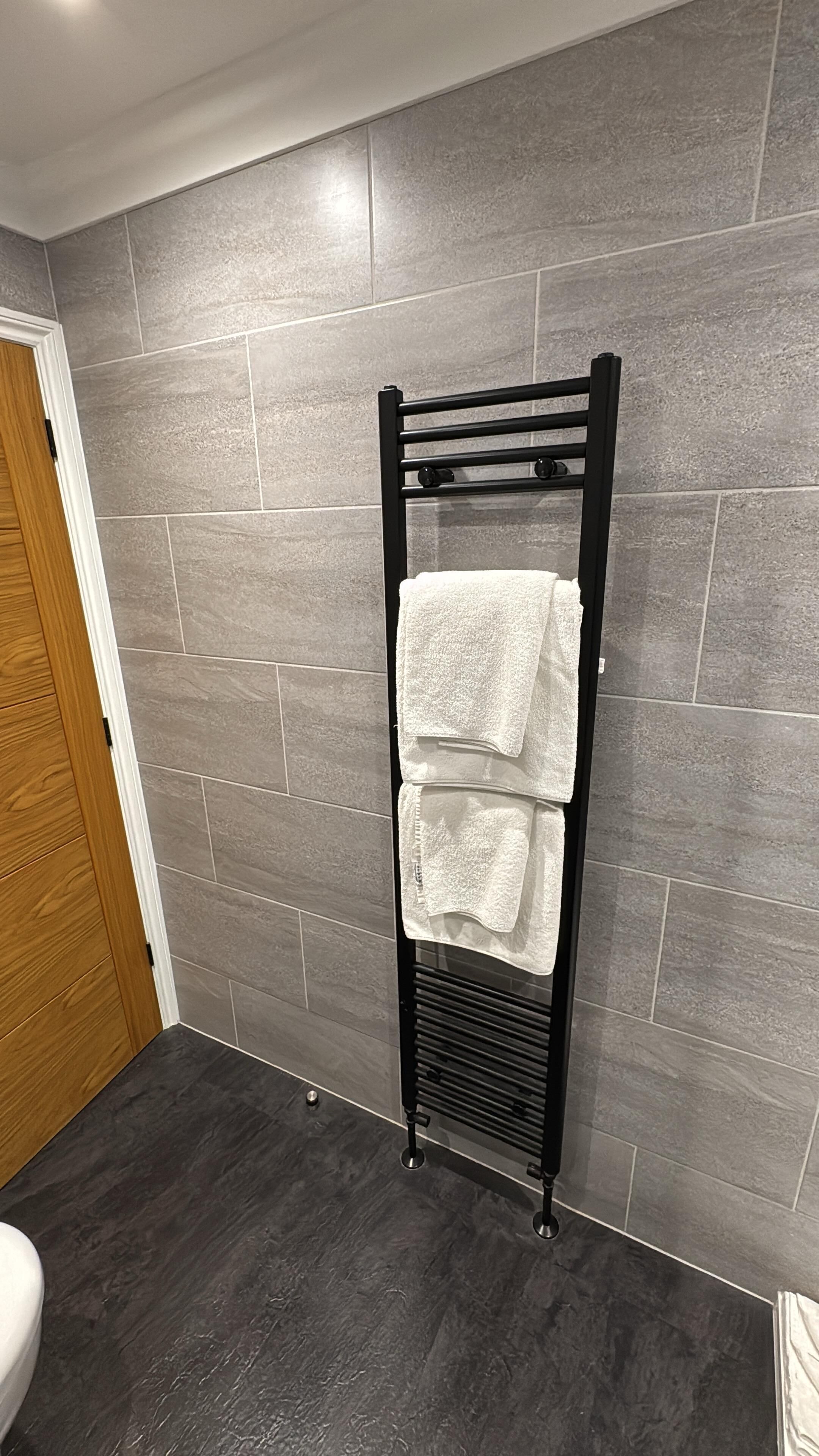 A bathroom with a black towel rack holding two white towels, grey tiled walls, and a wooden door.