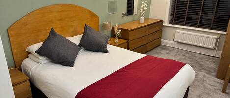 A neatly made bed with a wooden headboard, two grey pillows, and a red blanket. There is a wooden dresser with a vase of flowers on top, a bedside table with a lamp, and a window with blinds.