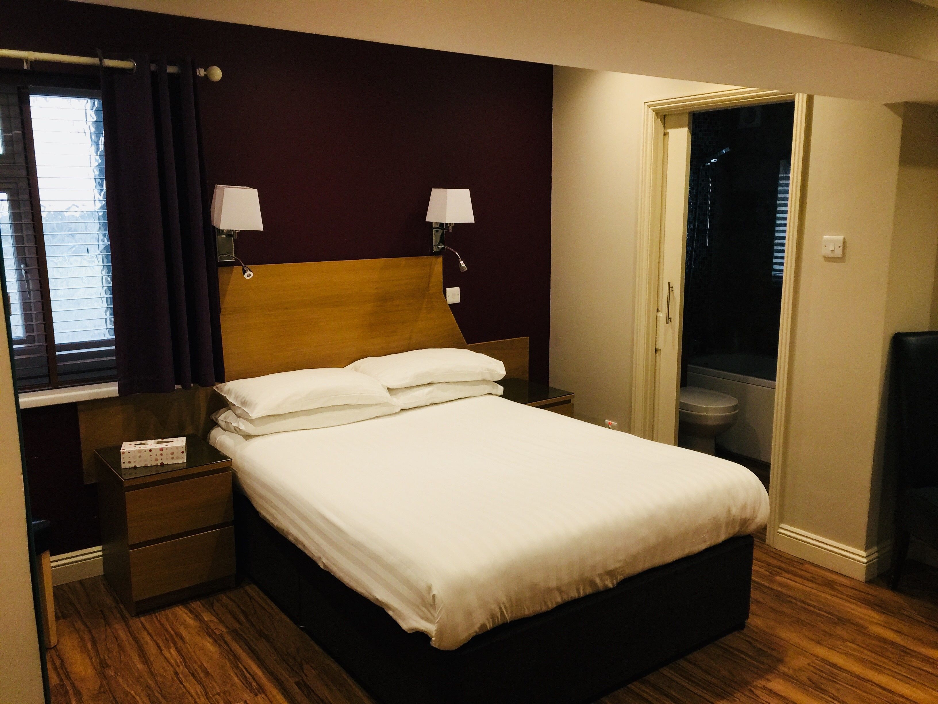 Una habitación de hotel con cabecera de madera, cama con sábanas blancas, dos lámparas de noche, una mesita de noche, una ventana con cortinas y una puerta que da a un baño.