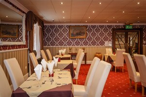 Comedor con mesas dispuestas para cuatro, sillas blancas tapizadas y papel pintado con estampado rojo.