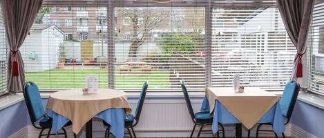 A dining area with two tables and four chairs, large windows with blinds, and a view of a residential area.