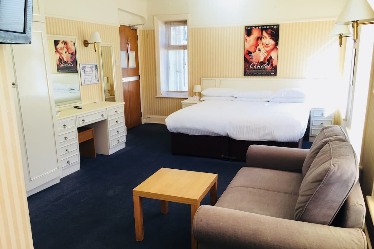 Hotellihuone, jossa on sänky, sohva, laatikoilla varustettu työpöytä, peili, ikkuna ja seinällä juliste.