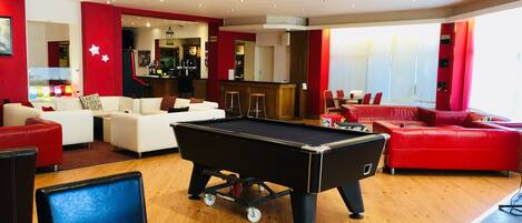 A pool table in a room with a red and white color scheme, a bar area, and a seating area with a red couch and white chairs.