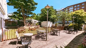 Ein Terrassenbereich im Freien mit Tischen und Stühlen, umgeben von Grünflächen und Gebäuden.