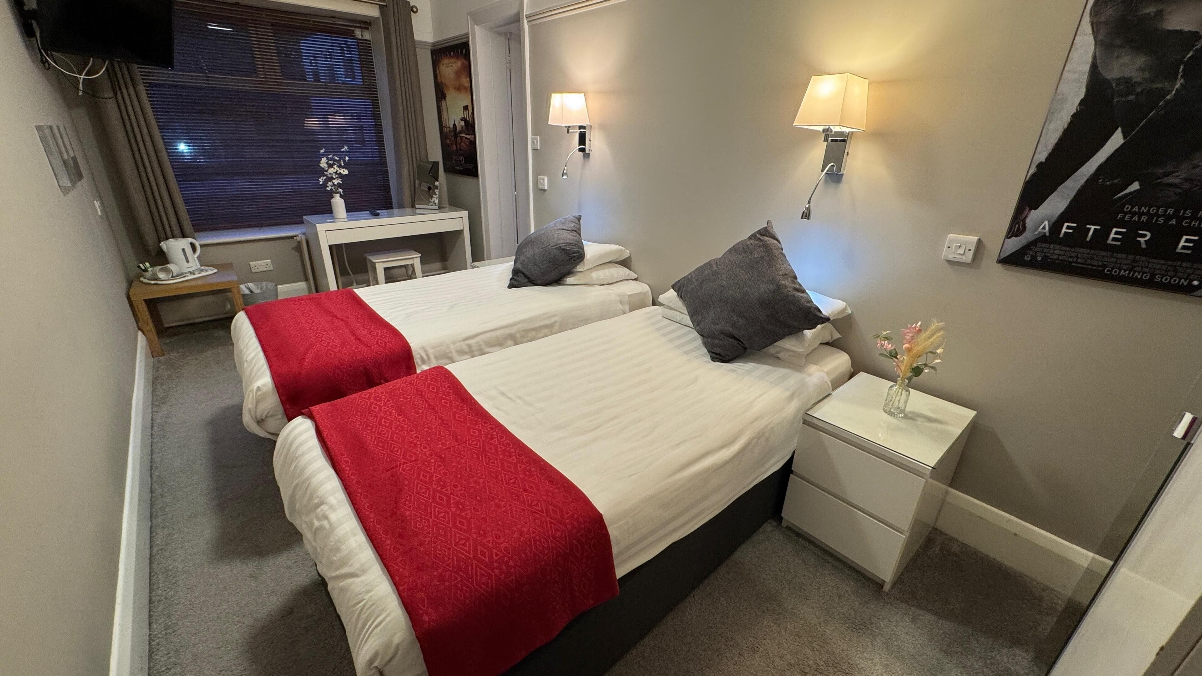 A hotel room with two beds, each with a red and white bedspread, a nightstand with a lamp, and a poster on the wall.