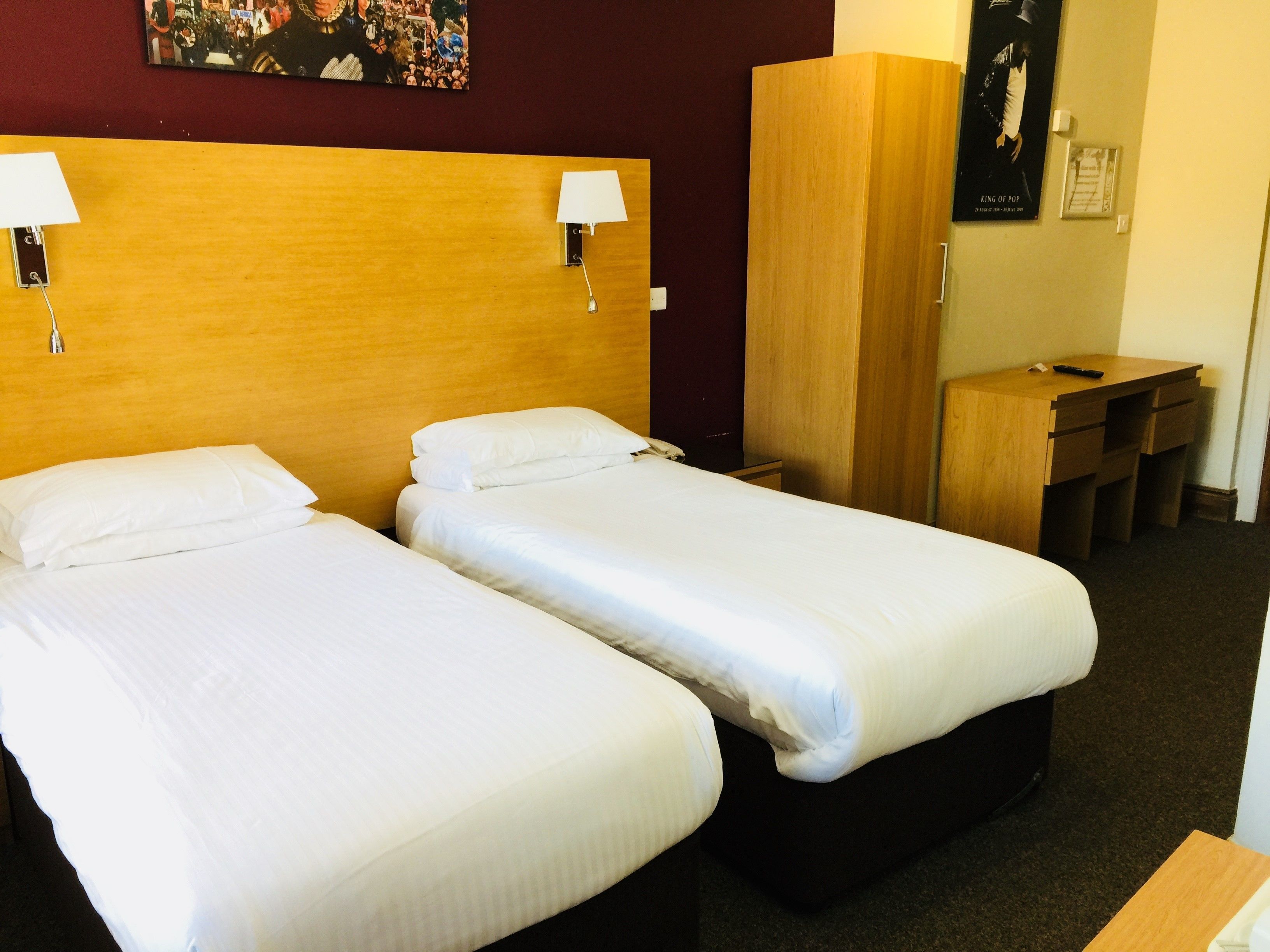 Ein Hotelzimmer mit zwei Einzelbetten, einem Holzschrank, einem Schreibtisch mit Stuhl und einem an der Wand befestigten Fernseher.