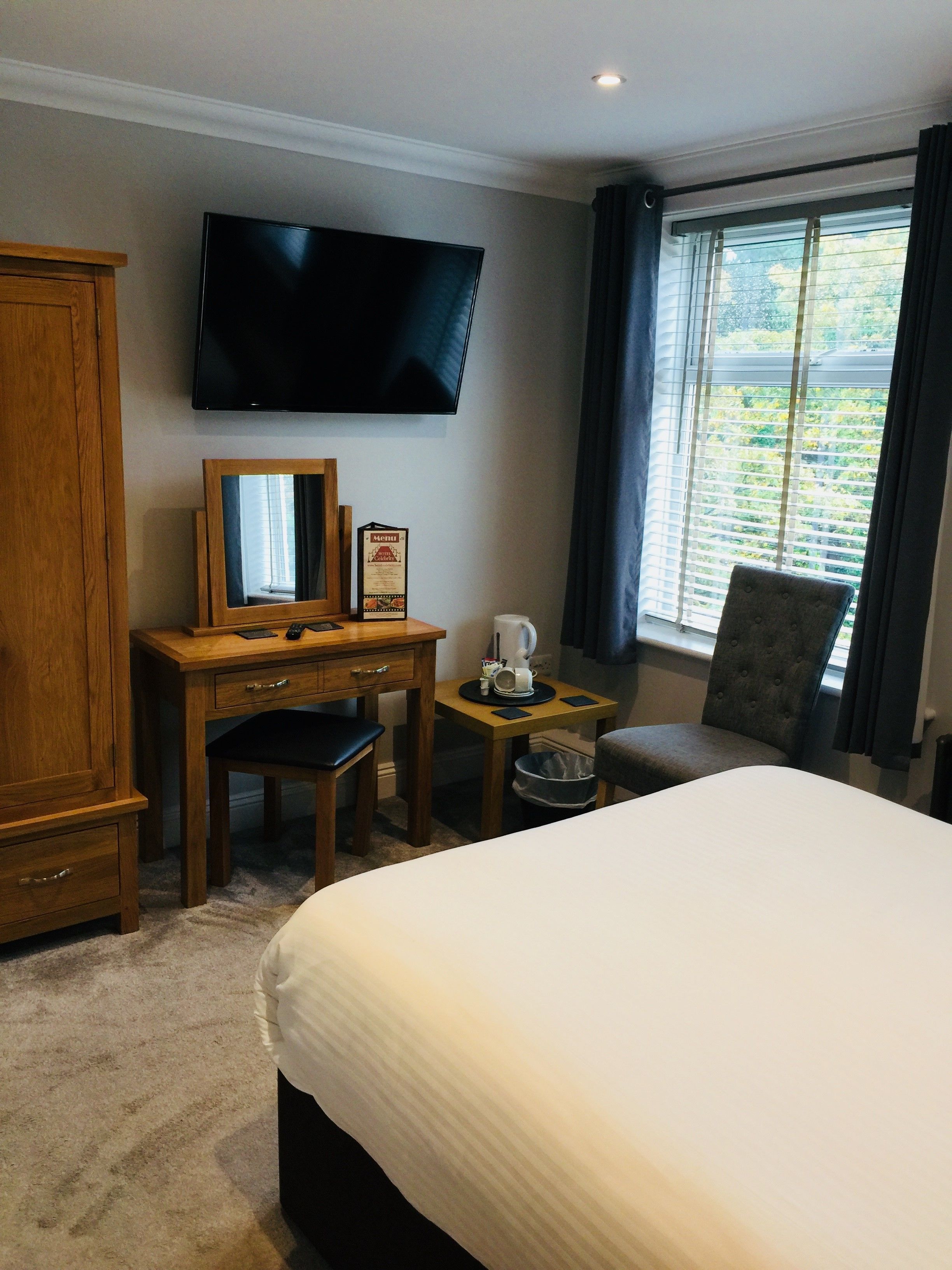 Een hotelkamer met een groot bed, een bureau met stoel, een aan de muur bevestigde televisie, een raam met gordijnen en uitzicht op groen.