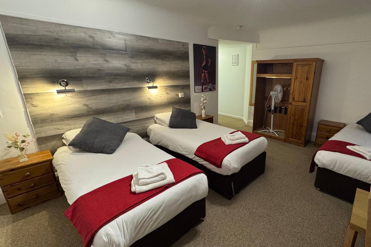 Hotellihuoneessa on kolme erillistä sänkyä, joissa kaikissa on punainen peitto ja valkoiset tyynyt. Huoneessa on myös puinen vaatekaappi, yöpöytä, jossa on lamppu, sekä seinään kiinnitetty valaisin.