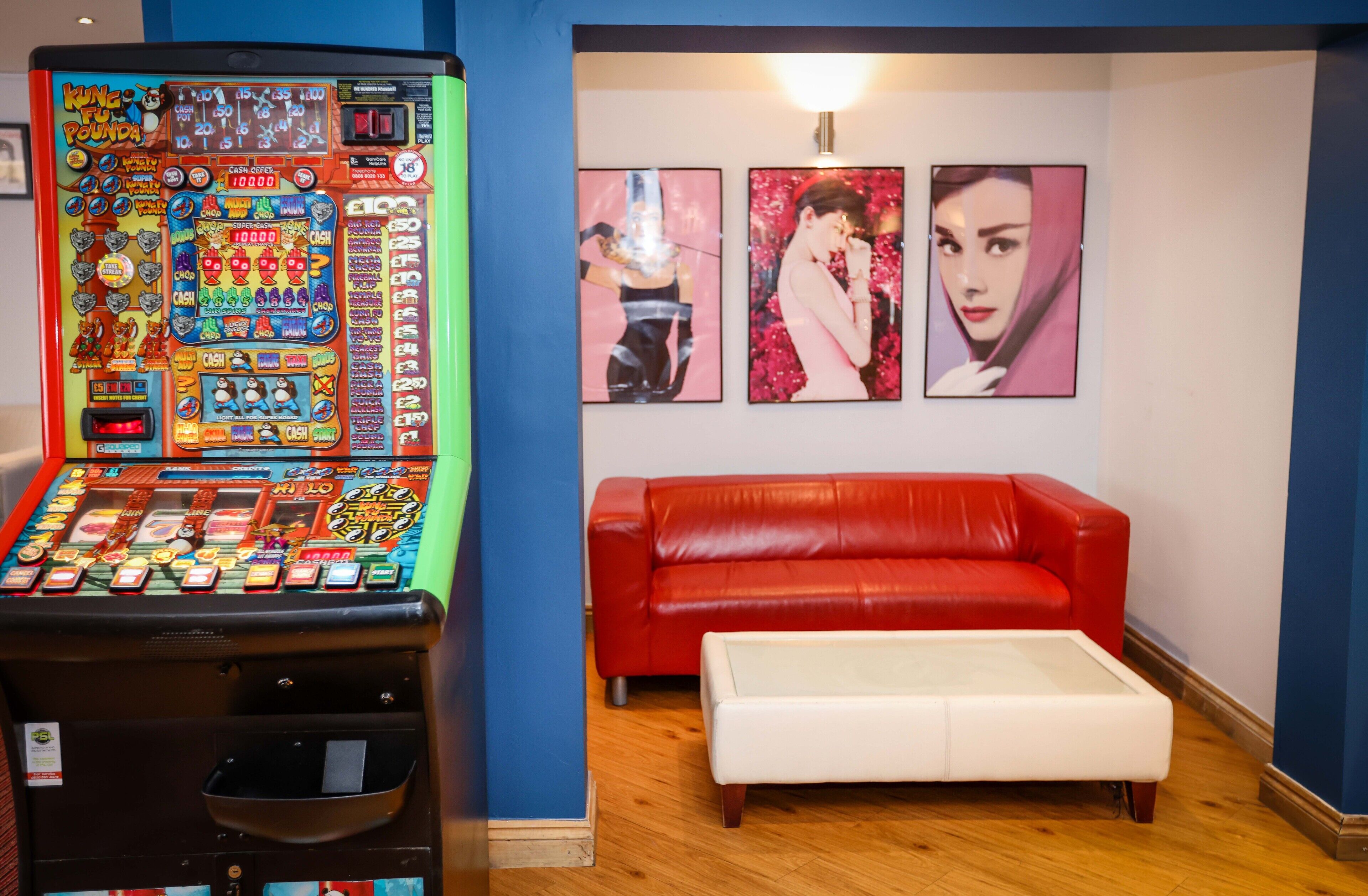 A room with a red leather sofa, a white ottoman, and a slot machine. There are three framed pictures on the wall.