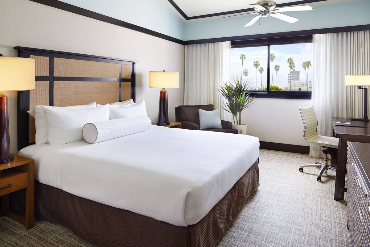 A hotel room with a large bed, two bedside tables, a desk, a chair, a ceiling fan, and a window with a view of palm trees.