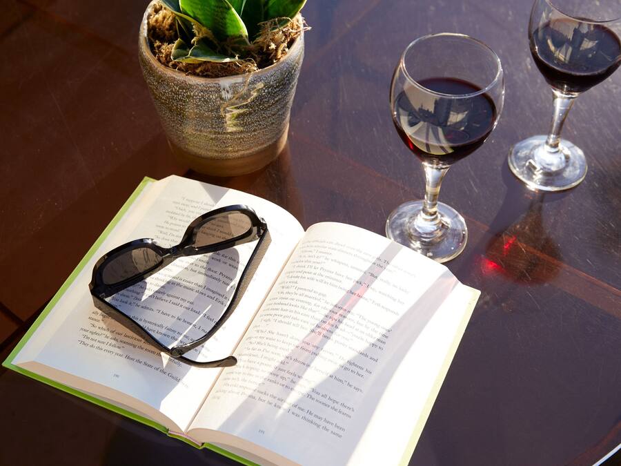 Un libro abierto con gafas encima, dos copas de vino con líquido rojo y una planta en maceta sobre una mesa.