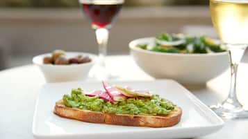 A plate with a slice of toast topped with guacamole and radish slices, a bowl of salad, and a glass of white wine on a table.