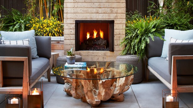 A modern outdoor seating area with a central glass coffee table, a wooden log fire pit, and surrounding plants. - The Ambrose (Santa Monica)