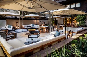 An outdoor patio area with multiple seating options including sofas, chairs, and tables. There are umbrellas providing shade, and the space is surrounded by plants and greenery.