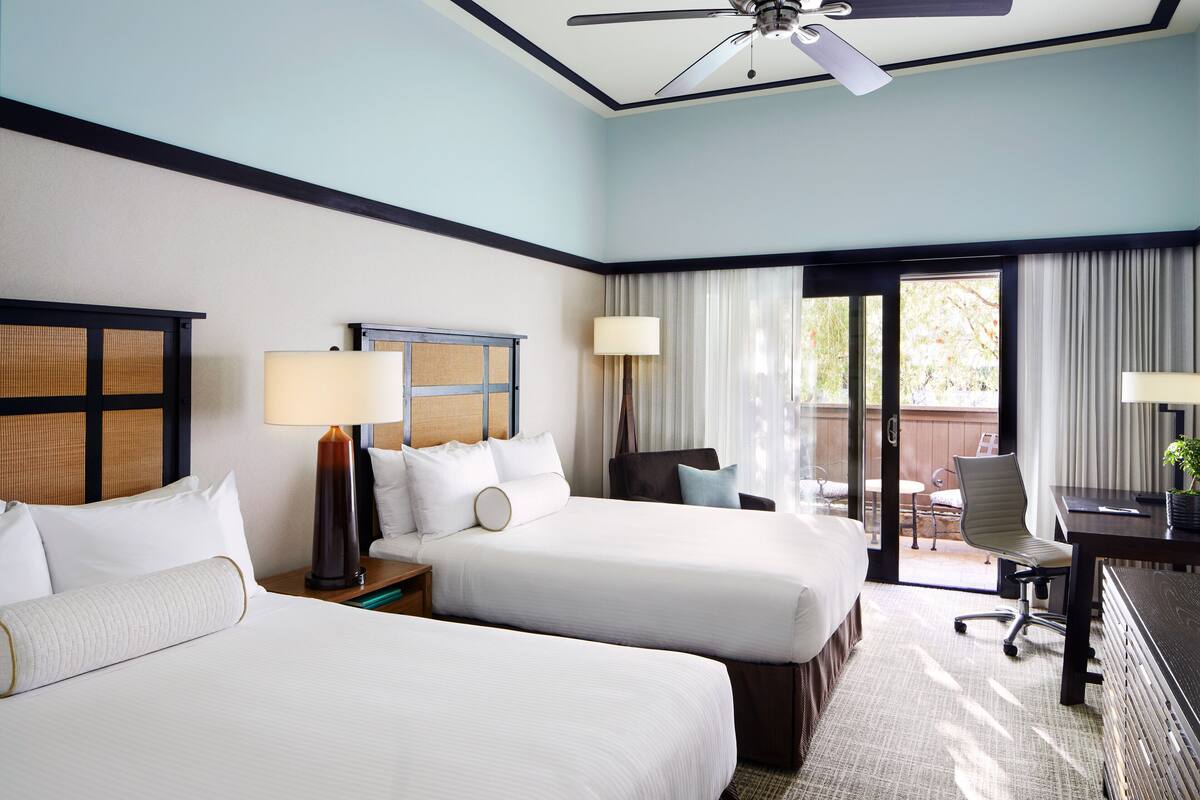A hotel room with two beds, a desk, a chair, a lamp, and a ceiling fan.