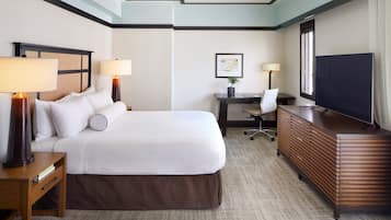 A hotel room with a large bed, two bedside tables with lamps, a desk with a chair, a television, and a framed picture on the wall.