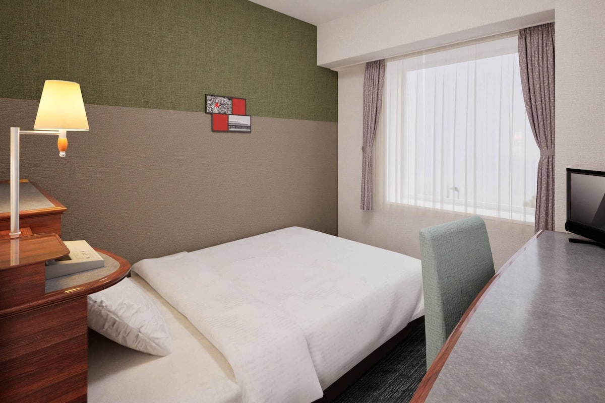 A hotel room with a bed, a desk with a lamp, a television, and a window with curtains.