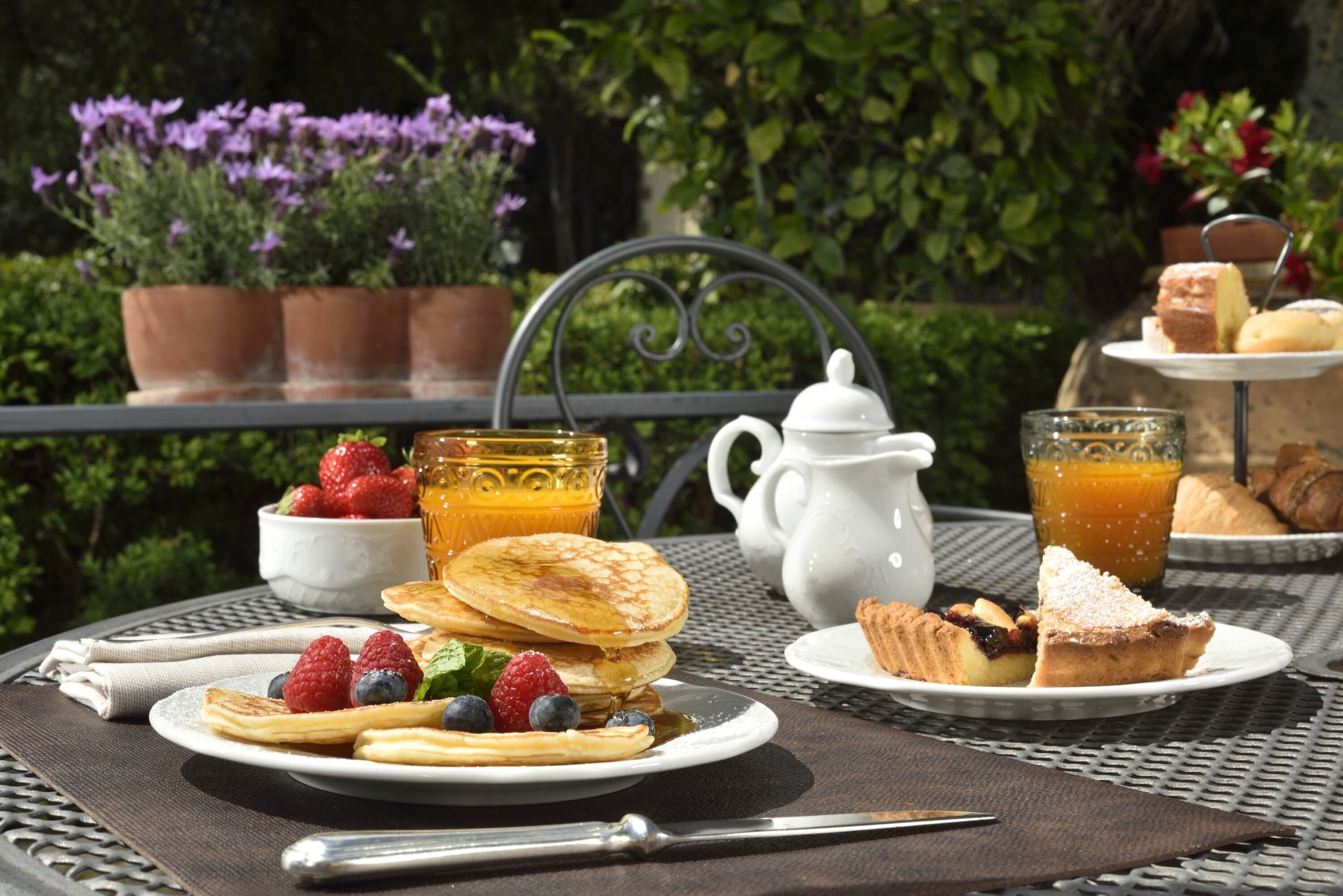 Una mesa de desayuno con panqueques, frutos rojos frescos, una jarrón, un vaso de jugo de naranja y pasteles.