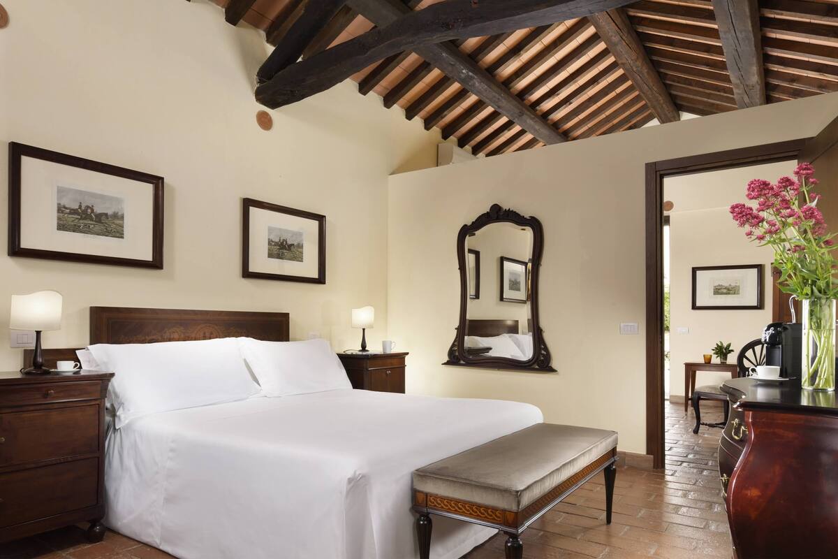A neatly made bed with a wooden headboard, a bedside table with a lamp, and a bench. There are framed pictures on the wall and a mirror above the bed.