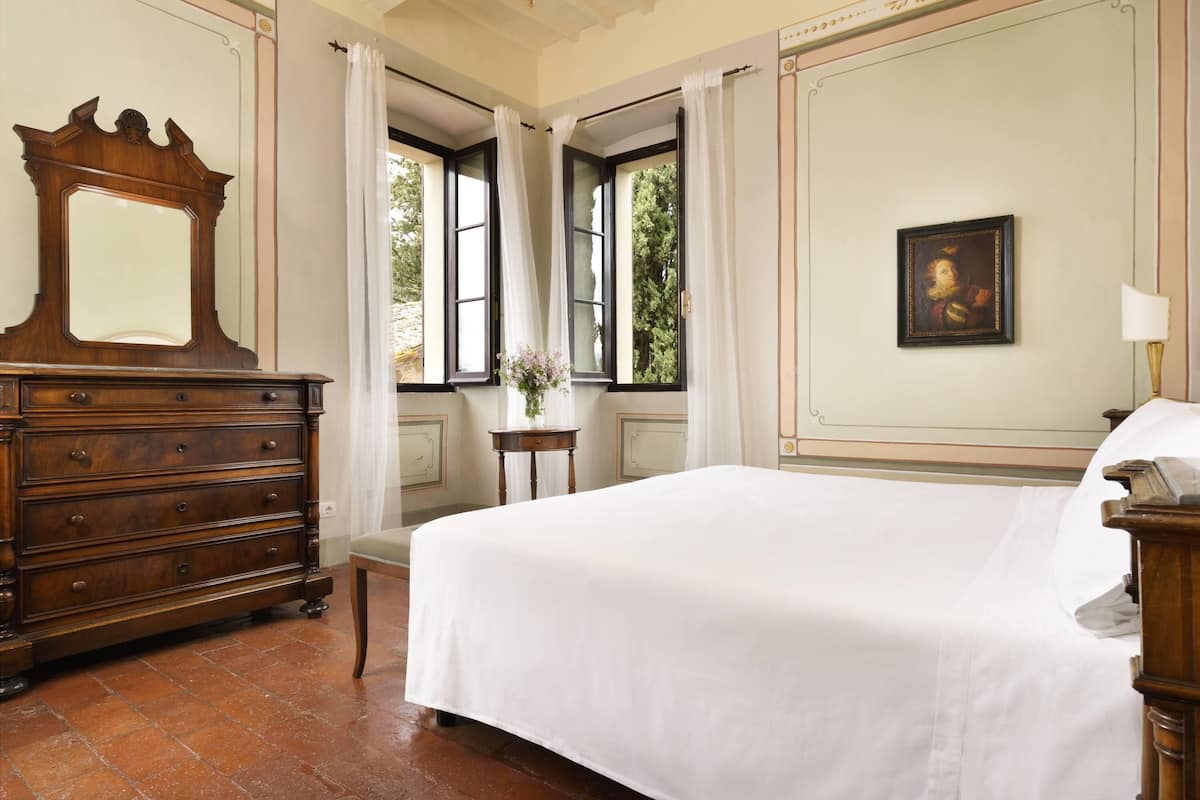 A bedroom with a large bed, a wooden dresser, a mirror, a window with curtains, and a painting on the wall.