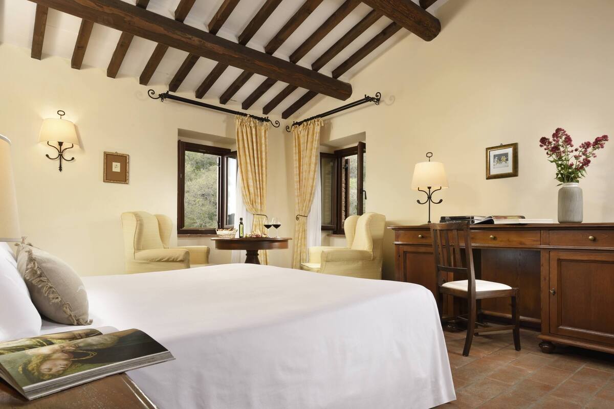A hotel room with a bed, two armchairs, a desk, a cabinet, and a vase with flowers.