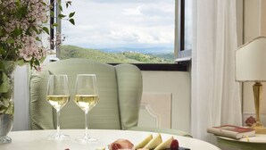 Una mesa de comedor con dos copas de vino blanco, una tabla de quesos con varios tipos de queso y una frambuesa, y una vista de colinas verdes a través de una ventana.