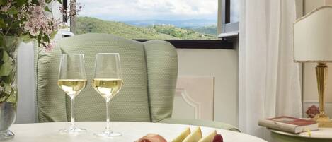 A dining table with two glasses of white wine, a cheese board with various cheeses and a raspberry, and a view of green hills through a window.