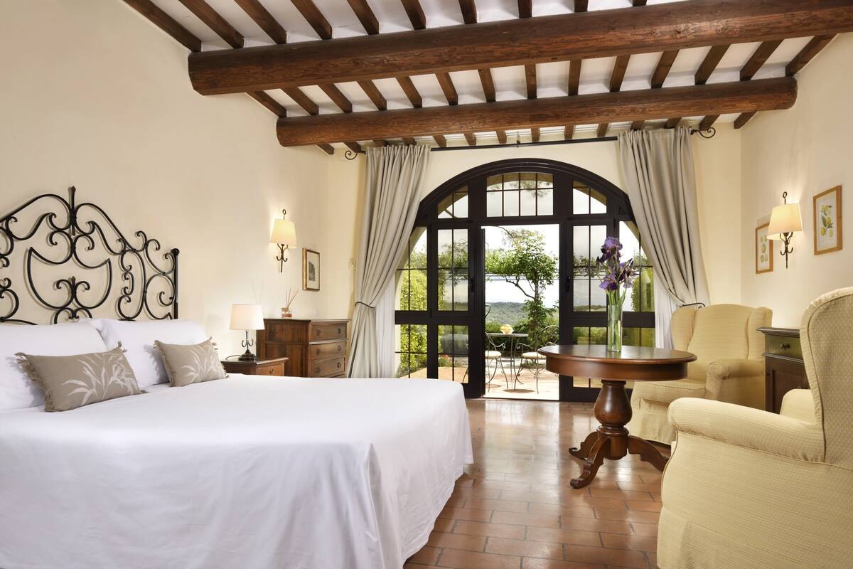 A bedroom with a large bed, a wooden ceiling, a table with chairs, and a view through an arched window.