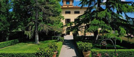 Un chemin menant à un bâtiment doté d’une tour, entouré de jardins et d’arbres bien entretenus.