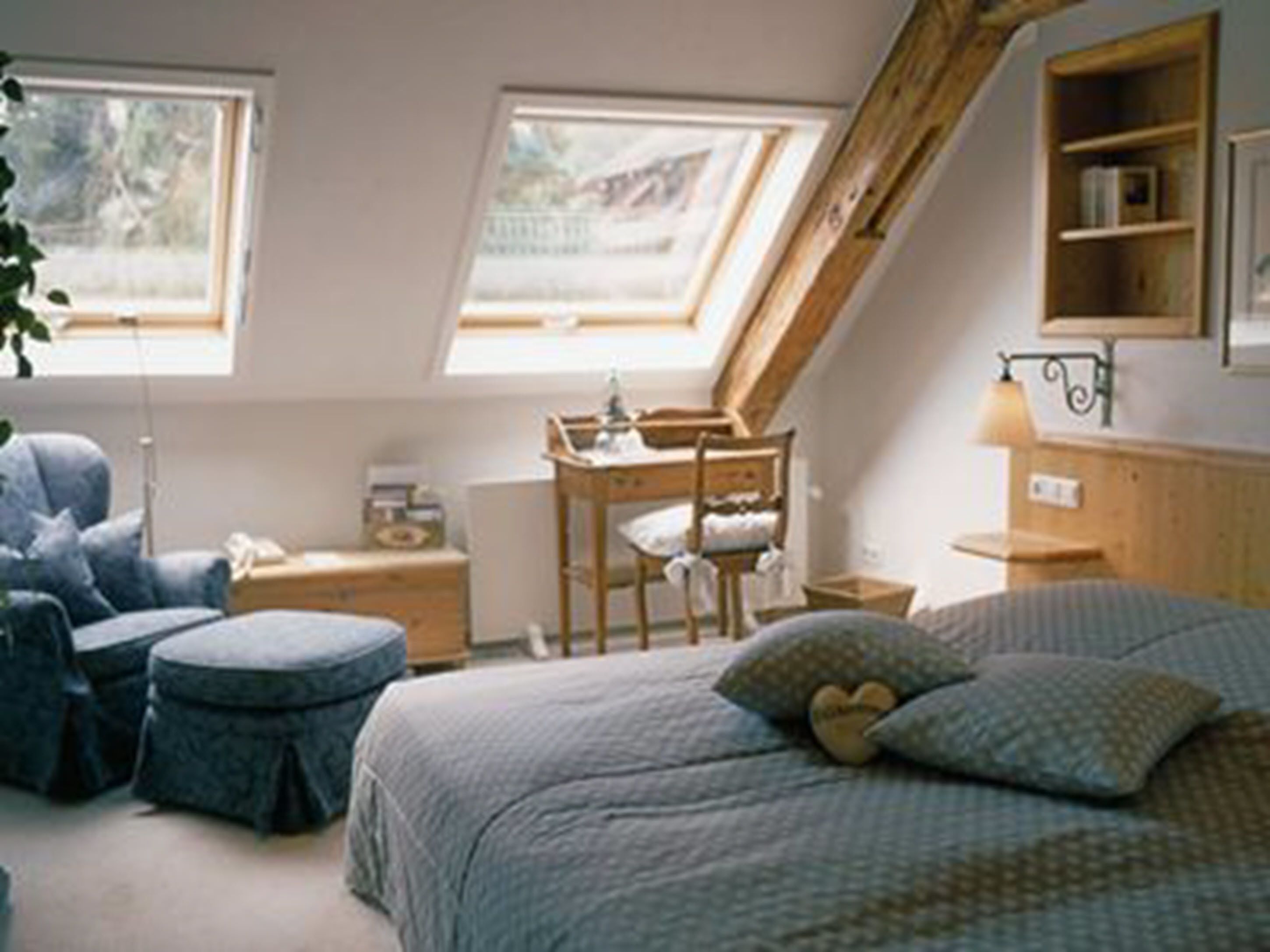 Une pièce chaleureuse avec un plafond en pente, un lit recouvert d’une couette à motifs, une chaise, une petite table et une étagère remplie de livres.
