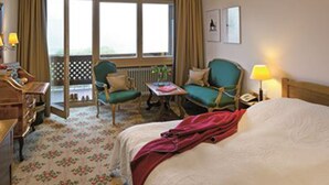 A hotel room with a bed, two chairs, a small table, a desk with a lamp, a window with curtains, and a view of the outdoors. - Romantik Hotel Spielweg (Münstertal)