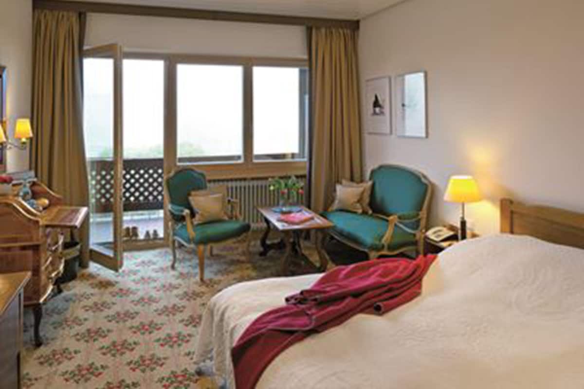 Een hotelkamer met een bed, twee stoelen, een kleine tafel, een bureau met een lamp, een raam met gordijnen en uitzicht op de omgeving.