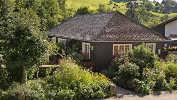 A small, dark-colored house with a sloped roof surrounded by lush greenery and a garden. - Romantik Hotel Spielweg (Münstertal)