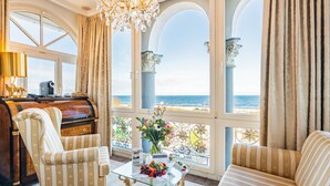 A luxurious room with a piano, striped armchairs, a glass coffee table with fruits, and a view of the sea through large windows. - SEETELHOTEL Ahlbecker Hof (Heringsdorf)