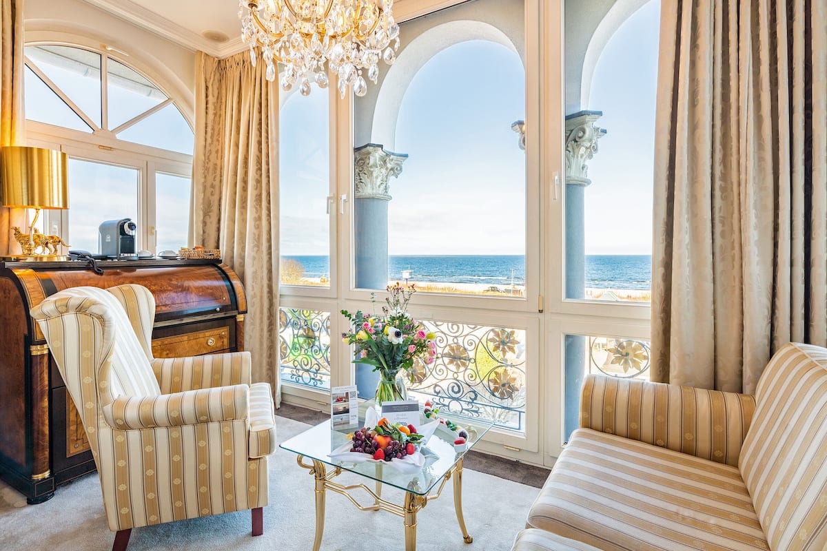 Une chambre luxueuse avec un piano, des fauteuils à rayures, une table basse en verre sur laquelle sont disposés des fruits, et une vue sur la mer par de grandes fenêtres.