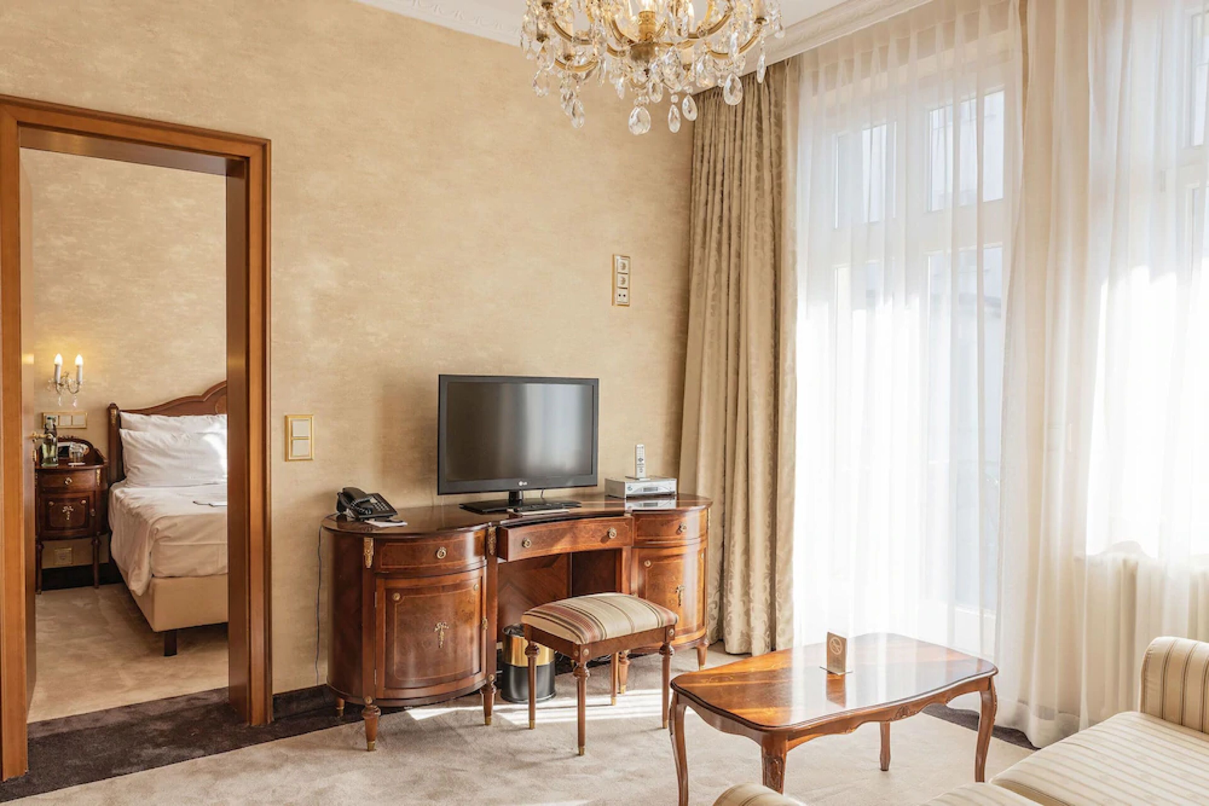 Una habitación de hotel con cama, escritorio con televisor, silla, mesa y un ventanal con cortinas.