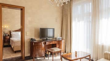 Una habitación de hotel con cama, escritorio con televisor, silla, mesa y un ventanal con cortinas.