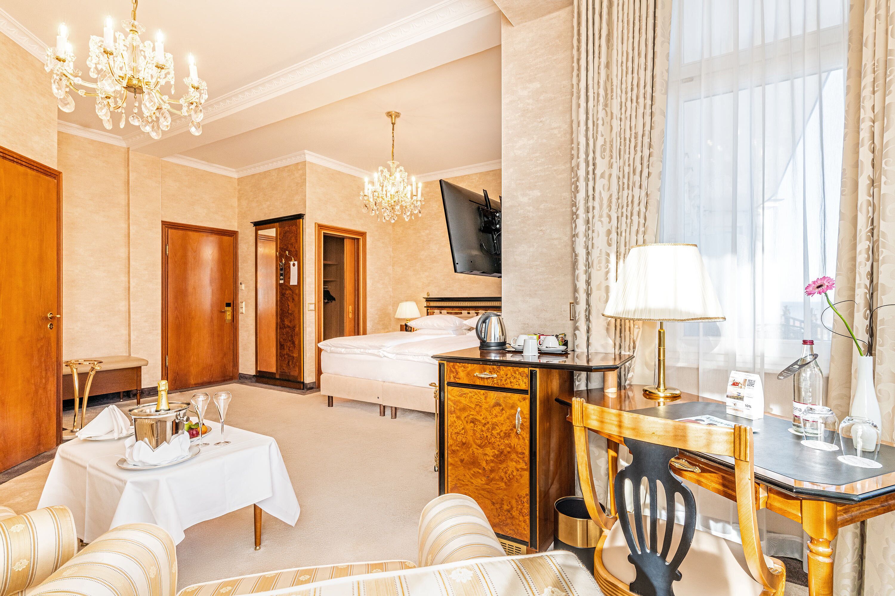 Habitación de hotel con cama, escritorio, silla, mesa con una botella y vasos, televisor y candelabro.