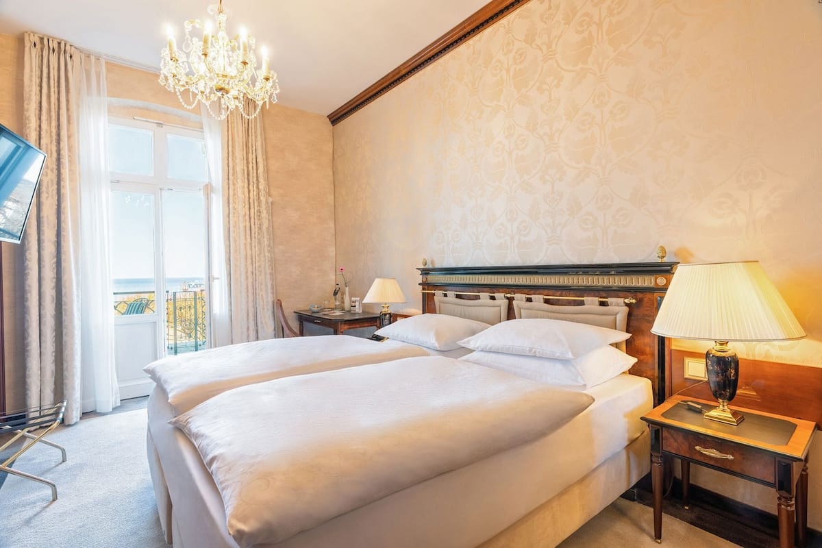 Une chambre d’hôtel avec un grand lit, un lustre, une table de chevet avec une lampe, un bureau et une vue sur l’extérieur.