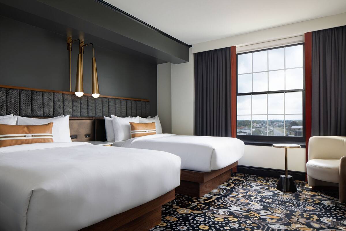 A hotel room with two beds, a small table, a chair, a large window with curtains, and a patterned carpet.