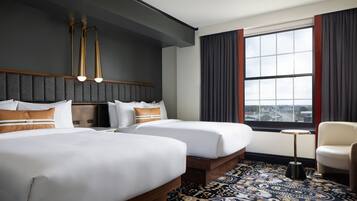 Habitación de hotel con dos camas, una mesita, una silla, un ventanal grande con cortinas y una alfombra estampada.