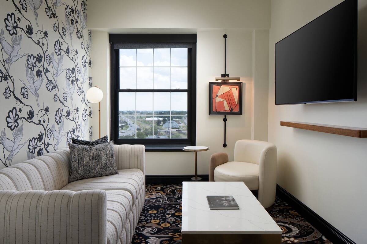 A modern hotel room with a large window, a sofa, a chair, a coffee table, and a flat-screen TV.