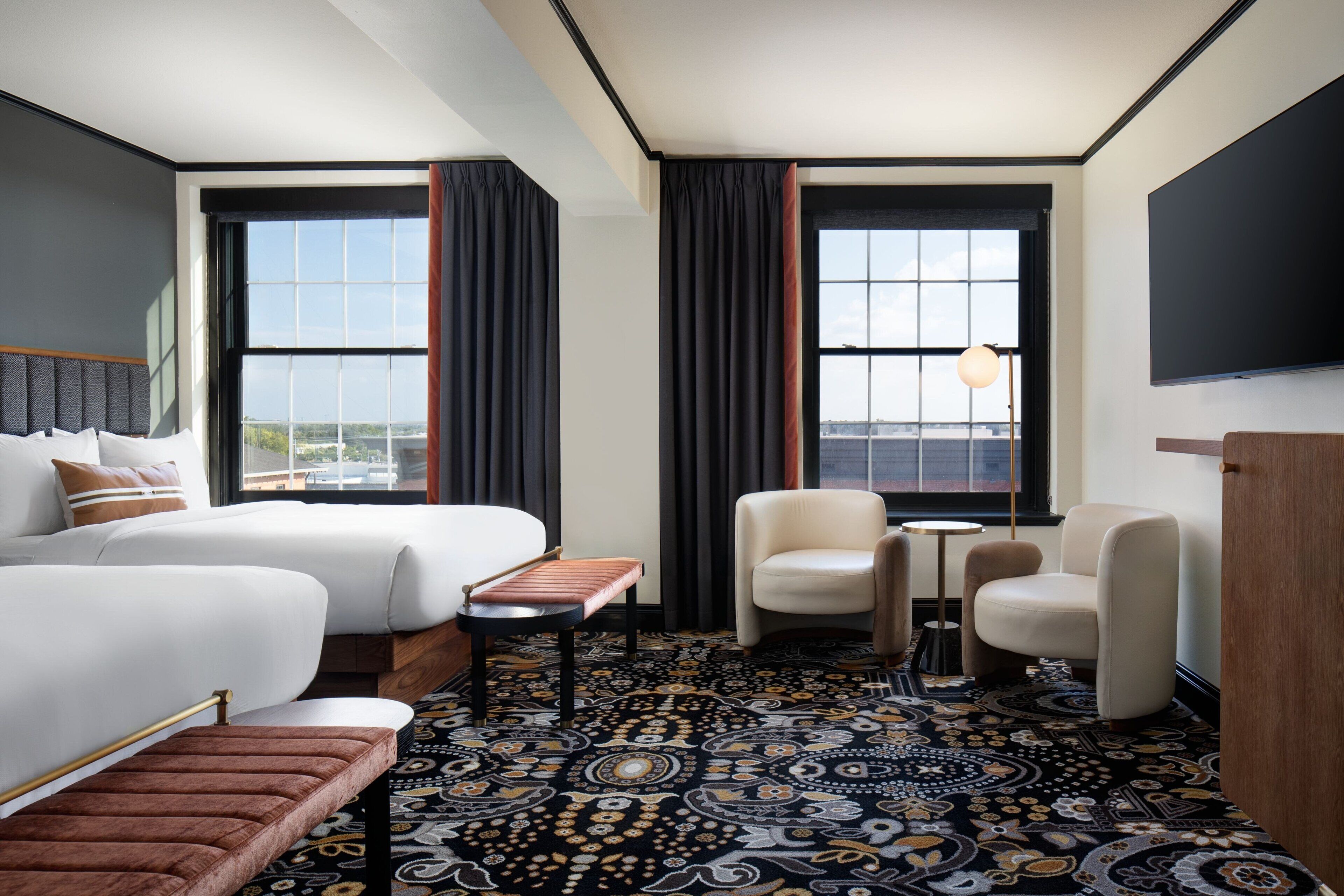 Habitación de hotel con dos camas, una mesita, dos sillas, un televisor y ventanas amplias con cortinas.