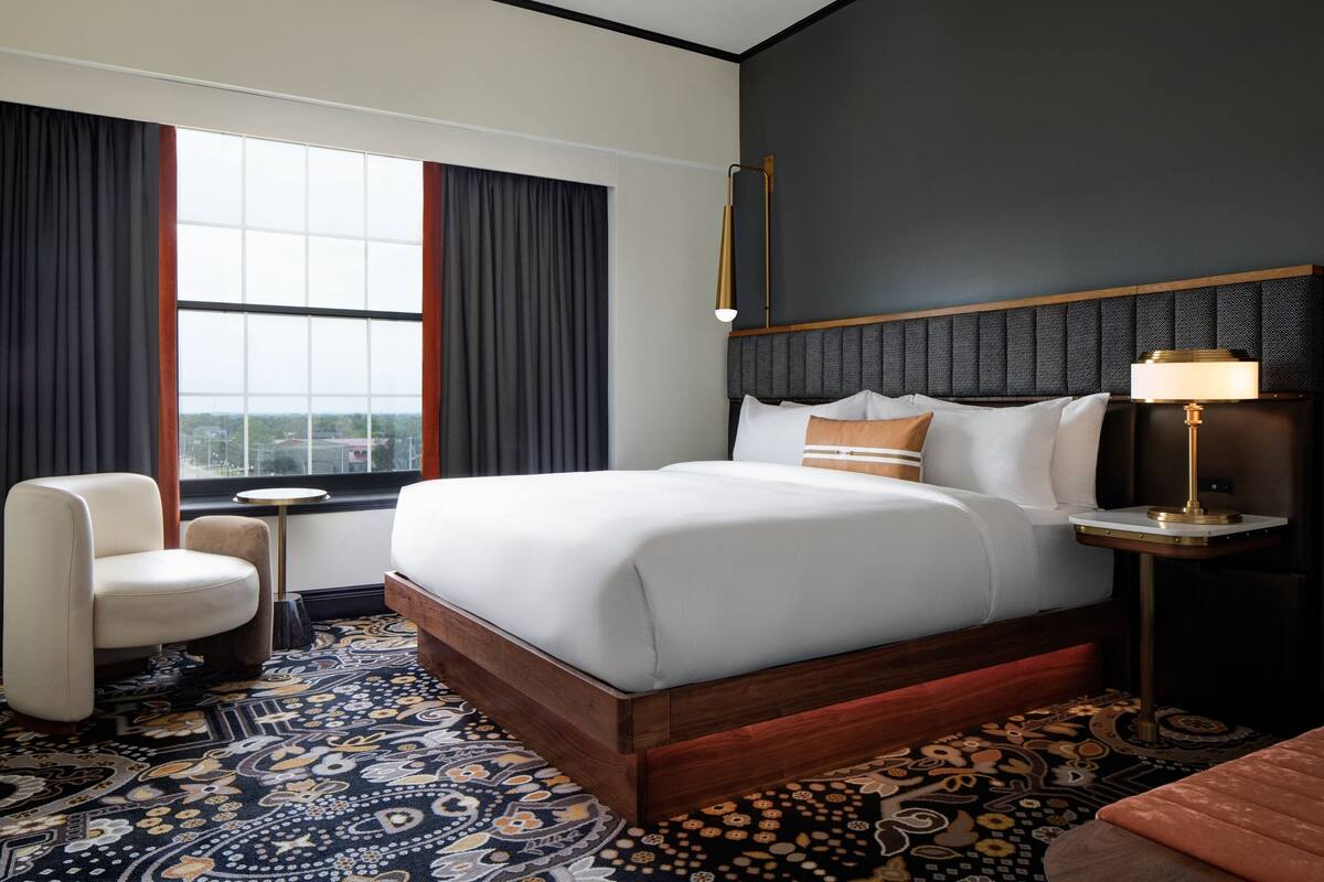 A hotel room with a large bed, a chair, a small table, and a lamp.