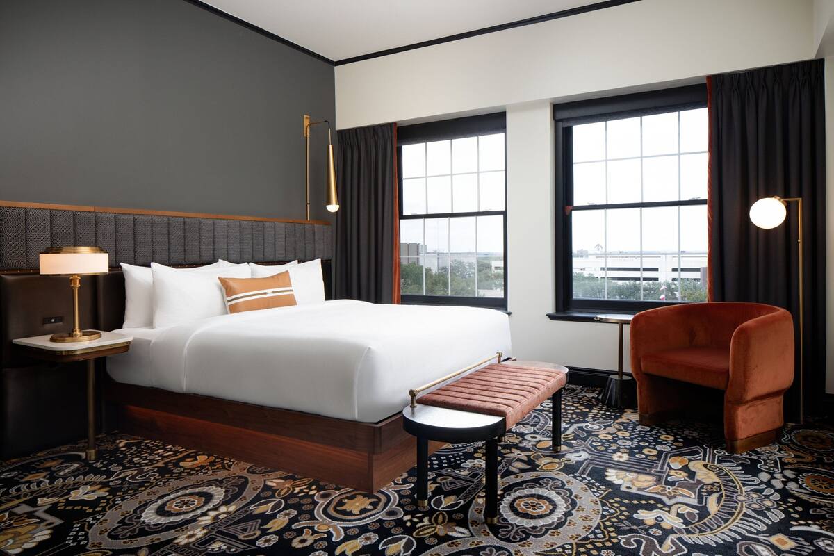 A hotel room with a large bed, a sitting area with a bench, a small table, and a chair. There are two windows with curtains, a patterned carpet, and a wall-mounted lamp.